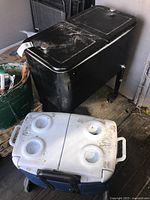 Blue Rubbermaid cooler with white lid showing four cup holders and dirt marks, positioned above large black table cooler.