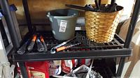 Photo showing garden hand tools on a plastic shelf including saw blade without handle, hand rake, cultivator, a plastic pot, woven basket, and partial containers below