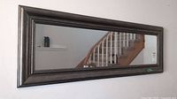 Clear rectangular mirror with dark wooden frame reflecting staircase and part of the room