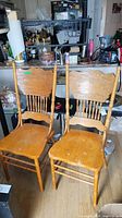 Image showing two wooden chairs with carved pressback and spindle details, light honey finish, placed indoors on wooden floor near kitchen counter.