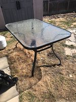 Image of rectangular outdoor patio table with a textured clear glass top and black metal frame standing on dry grassy ground outdoors.