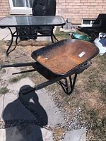 Full side view of the metal wheelbarrow on grass with patio table in background, showing overall shape and condition.