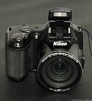 Front view of black Nikon COOLPIX L820 digital camera with flash popped up and textured grip visible.