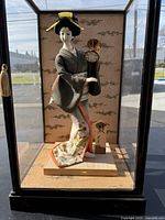 Full view of the Japanese Geisha doll inside glass display case showing the kimono, fan, hair accessories and wooden base.