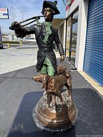 Front view of full statue outdoors on table showing hunter holding horn and two hounds with building in background
