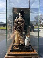 Geisha doll in traditional attire inside glass case with decorative vase and wooden base from front view