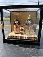 Photo of two antique Japanese geisha dolls and a small lantern displayed inside a glass display case with black frame, showing details of the dolls, clothing, and accessories.