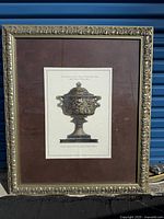 Framed lithograph showing ornate urn with Latin text, decorative frame and brown matte mounting