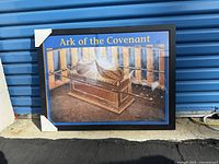 Framed poster of the Ark of the Covenant with black frame and corner protectors, placed outside against a blue garage door.