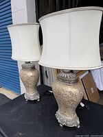 Pair of table lamps displayed outdoors on a black table next to a storefront. Visible are the white fabric shades and sparkling textured bases with silver-tone accents and feet.