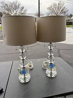 Pair of table lamps with clear glass spherical bases and beige drum lampshades, cords visible.
