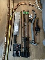 Wide view of all items arranged on wooden floor, showing bow, arrows, machete, walking stick, boomerang, and survival belt.