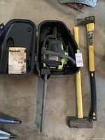 Photo of Poulan chainsaw inside black plastic carrying case with manual, Rockforge axe with yellow-black handle, and sledgehammer with yellow-black handle lying on floor.