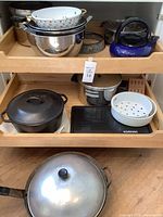 Overview of the cookware set on two wooden shelves including Lodge Dutch oven, enamel colanders, kettle, mixing bowls, wok, and other cookware items.