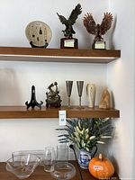 Shelf view with pottery plate, metal trophies, metal goblets, ceramic pumpkin, pottery figurines, clear glass and ceramic vases, and artificial flowers.