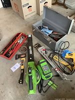 Wide layout of tools including metal toolbox, red tray with hand tools, Greenworks leaf blower with charger, small axe, hand tool set, Husky pipe wrench, and Dewalt corded drill on concrete floor.