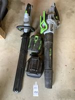 Photo of the EGO leaf blower, string trimmer, battery, and charger laid out together on a concrete floor showing condition and components.