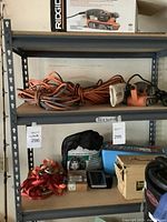 Shelf with orange extension cords and Ridgid belt sander with dust collection bag, along with tire chains, tie-downs, ammo box, and wooden clamps on lower shelf.