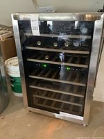 Frigidaire wine refrigerator in stainless steel with glass door, wooden shelves inside, displaying digital temperature readouts and several wine bottles.