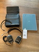 Harman/Kardon headphones laid out on a wooden surface alongside their carrying case and blue Apple iPad Smart Folio case in packaging.