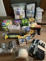Overview photograph of survival supplies including gloves, medical kits, light sticks, flashlights, paracord, disposable masks, and first aid items organized on floor.