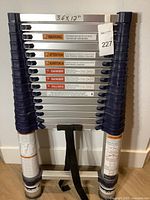 Photo of folded aluminum telescopic ladder showing multiple warning and usage labels and a tag with number 227.