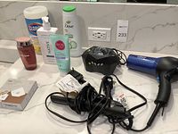 Photo showing assortment of personal care items including Clorox wipes, Dove lotion, Native deodorant, eos lip balm, and a red bottle. Also shows electronic devices including a black device with buttons, Chi hair straightener with cords, and a blue Conair hair dryer with cord on a white marble countertop.