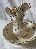 Round basin in background with scalloped rim decorated with floral and gold accents. Matching pitcher displayed inside basin showing ornate handle and spout with similar decoration.