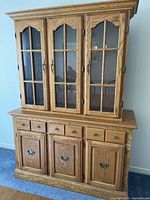 Front view showing solid oak cabinet with 3 drawers and 3 doors, topped by detachable hutch with 3 glass-paneled doors and brass fixtures.