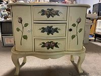 Front view of the cream wooden cabinet with floral designs on drawers and doors, showing its curved legs and scalloped base.