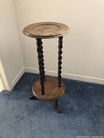 Full view of the decorative plant stand showing round table top and twisted spiral legs with a raised lip on top and base edges, finished in mahogany stain, set against a white wall and blue carpet.