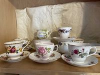 Photo showing 10 English bone china teacups and 9 saucers with assorted floral designs arranged on a shelf with a light cloth backdrop.