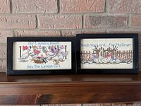 Two framed needlepoint artworks side by side on a wooden shelf against brick background, showing laundry room slogans with geese and laundry items in colorful thread on white fabric.