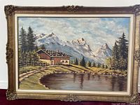 Full view of the oil painting showing alpine cottage near a lake with pine trees and mountains, inside an ornate wood frame