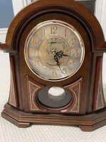 Front view of Seiko pendulum mantle clock showing wood casing, decorative dial with gold numbers and clock hands, and pendulum window.