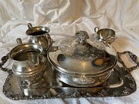 Set of five silver plated serveware items on a fabric background showing covered casserole, tray, two sauce boats, and creamer