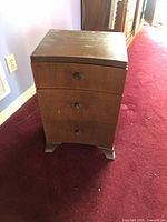 Front view of small brown wooden bedside table showing three drawers with round knob handles and short legs.