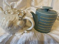 Side view of white strawberry tea pot and blue coffee pot placed next to each other on a wrinkled fabric.