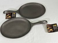 Top view showing two new Lodge OS2 cast iron griddle skillets with product tags attached, oval-shaped, black cast iron surface, single handle each.