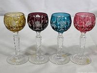 Full set of four mini cordial glasses in yellow, purple, blue, and pink colors with cut to clear design and clear faceted stems arranged in a row.