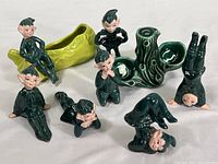 Full view of 8 vintage ceramic pixie figurines with green outfits and black hair, plus two green pottery log pieces