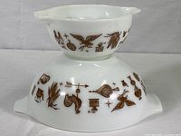 Two Pyrex Cinderella mixing bowls stacked, white milk glass with brown Early American pattern featuring eagle, rooster, coffee grinder, corn stalks, and other farm imagery.