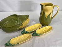 Photo of vintage Shawnee pottery covered serving bowl, 8-inch Shawnee pitcher, and three corn-shaped dishes from Walker Potteries arranged on flat surface.