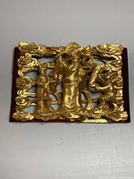 Front view of gold coloured carved wood hanging plaque showing three figures and floral/cloud motifs on reddish-brown wooden base.