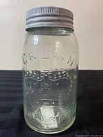 Front view of clear glass mason jar with embossed 'CROWN' and crown emblem with metal lid.
