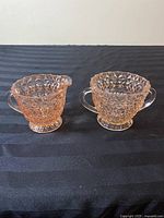 Photo of the front side of the pink pressed glass creamer and sugar bowl set showing textured diamond and ribbons pattern, spout on creamer, both with handles.