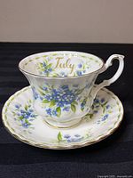 White bone china teacup with blue forget-me-not flowers, gold trim, and 'July' inscribed inside.