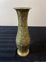 Front view of a solid brass tall vase with etched leaf and floral patterns on a black striped surface