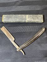 Photo of The Princes Own straight razor unfolded on black fabric with original case above.