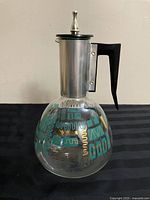 Front angle of vintage glass coffee carafe with stainless steel top and black handle, showing teal and gold mid-century geometric patterns.